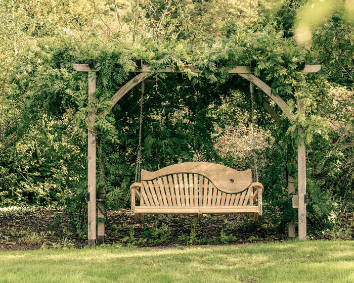 A wooden swing seat hangs beneath a leafy arbor surrounded by greenery.