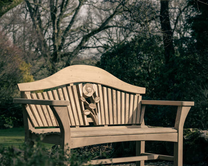 Wooden bench with a carved rose design, surrounded by trees and greenery.