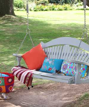 A painted swing bench hanging from a tree, with cushions and a picnic basket