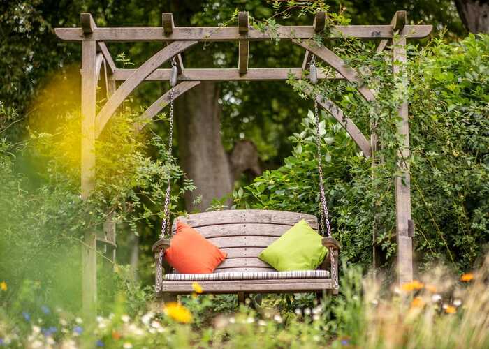 A pergola swing seat with green and orange cushions, surrounded by pretty greenery
