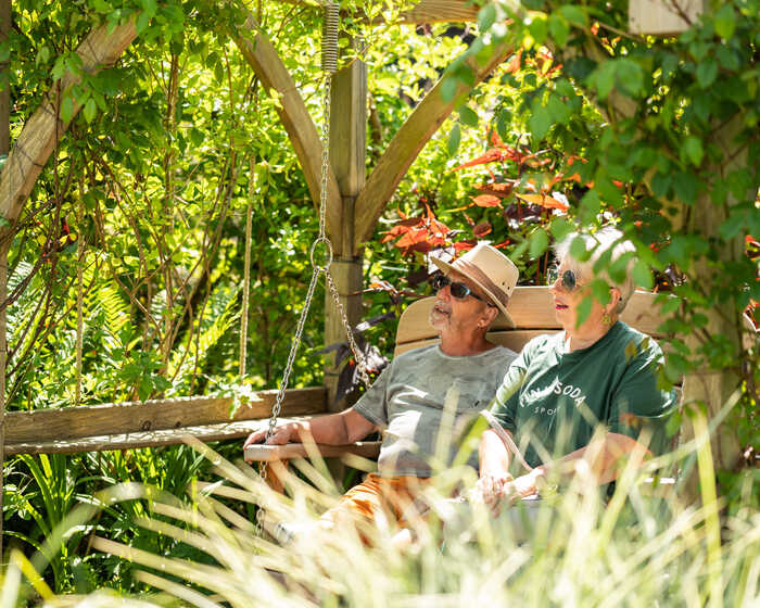 Two people relaxing on a swing seat and pergola in a lush garden, surrounded by greenery.